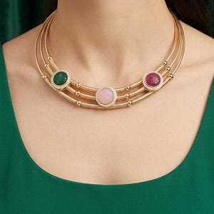 May include: Gold-toned necklace with three strands, each adorned with a central gemstone. The gemstones are green, pink, and red, each surrounded by a halo of small, clear stones. The necklace is worn against a green background.