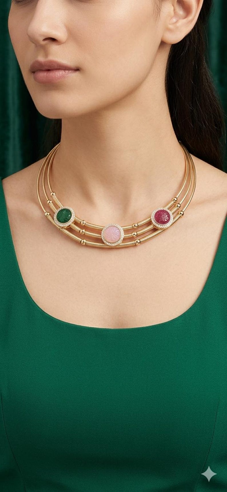 May include: Gold-toned necklace with three strands, each adorned with a central gemstone. The gemstones are green, pink, and red, each surrounded by a halo of small, clear stones. The necklace is worn against a green background.
