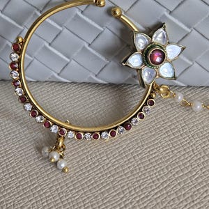 May include: A gold-toned circular nose ring featuring a flower-shaped accent with white petals and a central red stone. The hoop is embellished with alternating small white and red stones along the outer edge, with a small cluster of white beads.