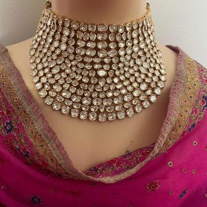 May include: A gold choker necklace set with numerous clear, oval-shaped stones. The necklace is broad and sits close to the neck. The background includes a pink and patterned fabric.
