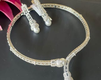 May include: A silver-toned necklace and earring set. The necklace is a choker style with a central pendant and a dangling pearl. The earrings are long, with a pearl at the bottom. The set is embellished with small, sparkling stones.