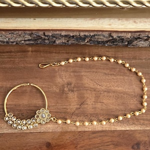 May include: A gold-toned nose ring with a floral design and clear gemstones. The ring is connected to a chain of small, round, cream-colored beads. The jewelry is displayed on a wooden surface.