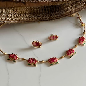 May include: Gold-coloured necklace and matching earrings set featuring pink lotus flower pendants with green accents. The jewellery is displayed on a white marble surface with a gold fabric in the background.