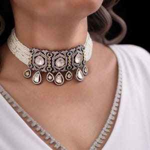 May include: A pearl choker necklace with a decorative centerpiece and teardrop pendants. The centerpiece features clear, faceted stones set in a silver and gold filigree design. The necklace is worn against a white top.