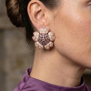 May include: A large flower-shaped stud earring featuring a central cluster of round mauve-colored stones surrounded by cream-colored petals with intricate gold-toned filigree detailing and a metallic gold-toned border along the outer edges.