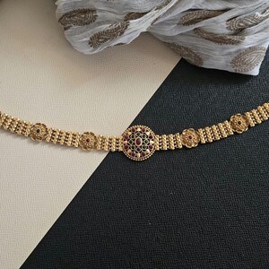 May include: Gold-coloured bracelet with a central circular design and floral accents. The centre features a cluster of red gemstones. The bracelet is set against a black and white background, with patterned fabric in the background.
