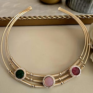 May include: A gold-toned collar necklace with three parallel bands. The necklace features three circular gemstone pendants: green, pink, and red, each encircled with small, clear crystals. The gemstones have carved floral designs.