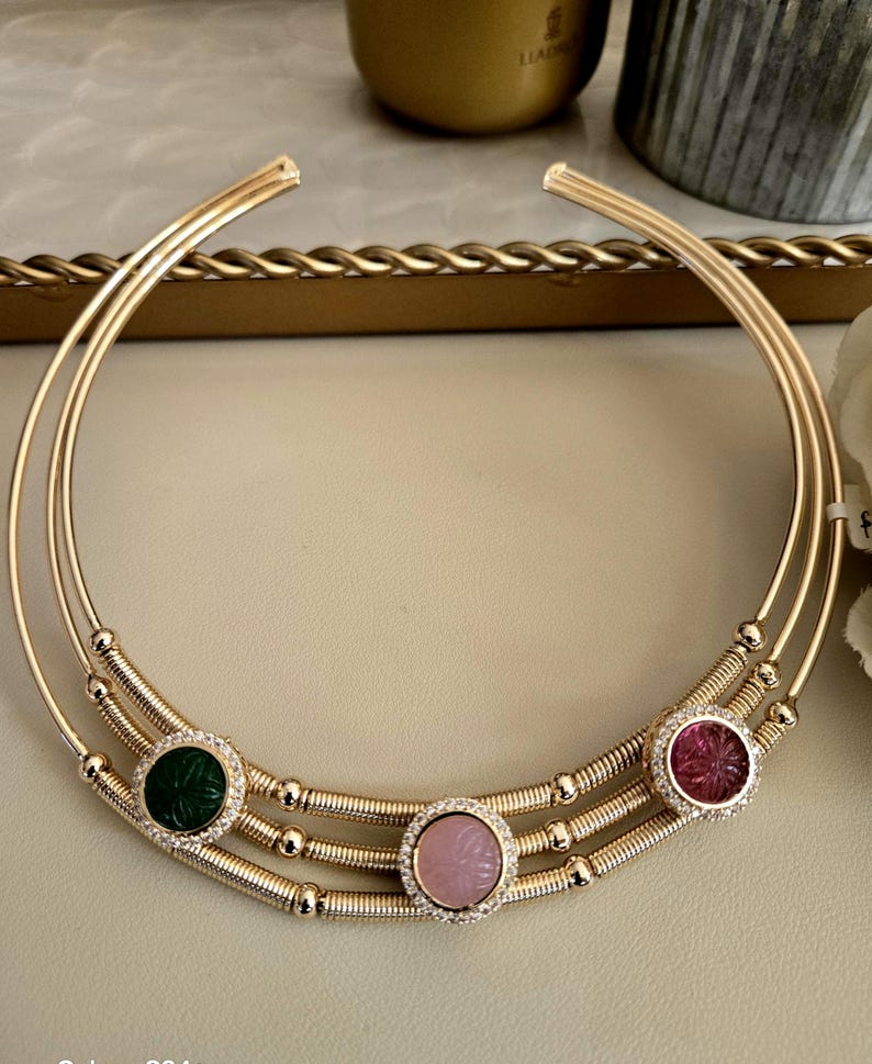 May include: A gold-toned collar necklace with three parallel bands. The necklace features three circular gemstone pendants: green, pink, and red, each encircled with small, clear crystals. The gemstones have carved floral designs.