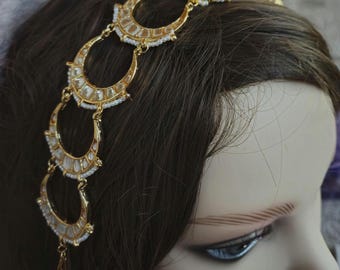 May include: Gold headband with a row of clear stones and white beads. The headband is decorated with a series of crescent moon shapes.