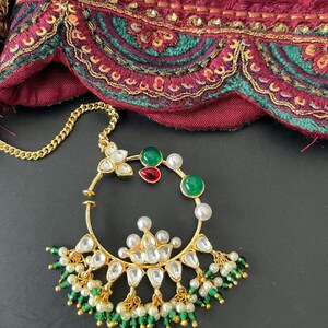 May include: A gold-toned nose ring with a floral design and dangling green and white beads. The ring features clear, green, and red gemstones, and a delicate gold chain. The background includes a maroon fabric with gold and teal embroidery.