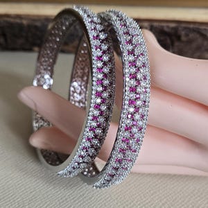 May include: Two silver bangles, each adorned with a repeating pattern of small, round, pink and clear stones. The bangles are displayed on a light pink surface. The jewellery is likely made of metal and gemstones.