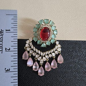 May include: An ornate earring with a large red oval center stone, surrounded by light green stones. Below, a crescent of clear stones and dangling pink teardrop gems. The earring is approximately 2 inches in length.