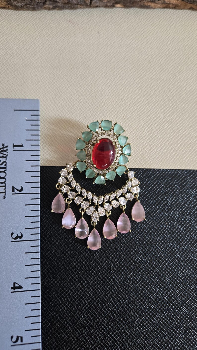 May include: An ornate earring with a large red oval center stone, surrounded by light green stones. Below, a crescent of clear stones and dangling pink teardrop gems. The earring is approximately 2 inches in length.
