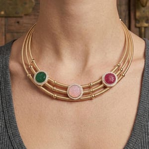 May include: A gold necklace with three strands, each adorned with a gemstone pendant. The pendants are green, pink, and red, each surrounded by small clear stones. The necklace is displayed on a person wearing a gray top.