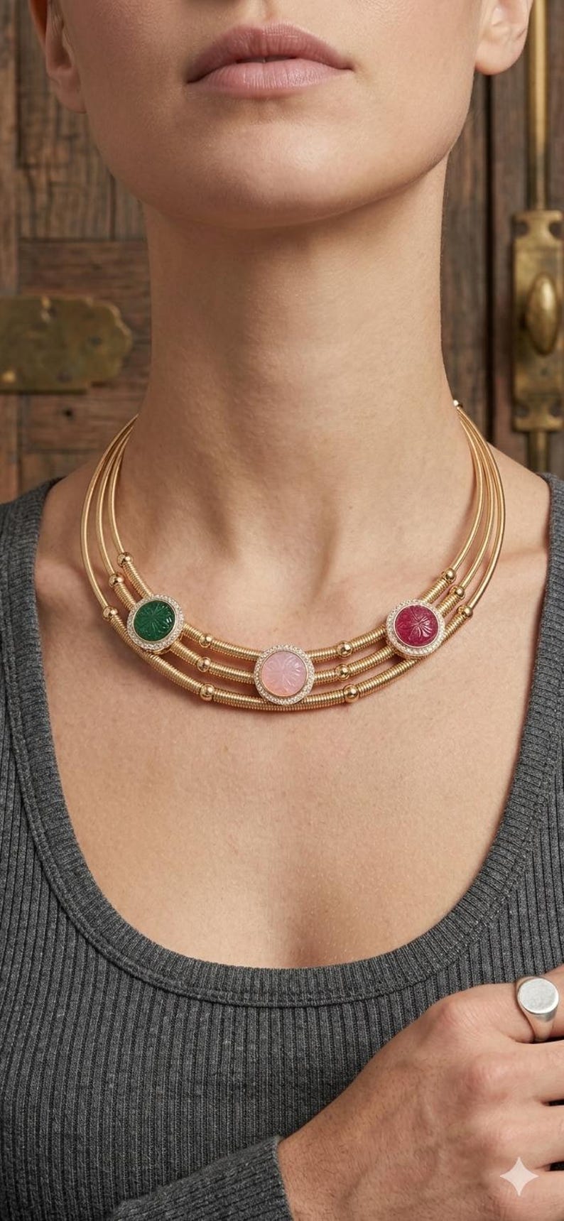 May include: A gold necklace with three strands, each adorned with a gemstone pendant. The pendants are green, pink, and red, each surrounded by small clear stones. The necklace is displayed on a person wearing a gray top.