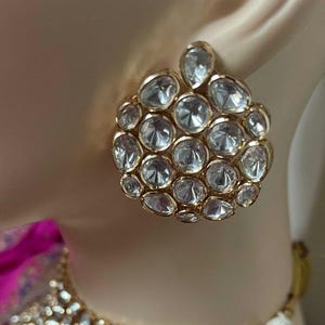 May include: Gold-coloured stud earrings with a cluster of clear, faceted stones. The earrings are round with a smaller, teardrop-shaped cluster at the top. The stones are set closely together, creating a sparkling effect.