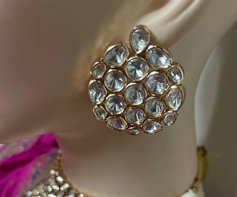 May include: Gold-coloured stud earrings with a cluster of clear, faceted stones. The earrings are round with a smaller, teardrop-shaped cluster at the top. The stones are set closely together, creating a sparkling effect.