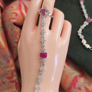 May include: A silver-toned hand chain featuring a ring connected to a bracelet by a central strand. The jewelry is adorned with clear white stones and rectangular and oval-shaped magenta-colored stones set in a metallic frame with intricate detailing.