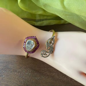 May include: Two gold-toned bracelets. One features a large, clear, faceted stone surrounded by small, red stones. The other bracelet has a decorative, openwork design with small, clear stones. The bracelets are on a light-skinned arm.