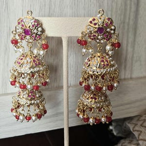 Peut inclure: Boucles d'oreilles pendantes dorées à trois niveaux. Les boucles d'oreilles présentent des accents rouges et blancs, avec un dessus floral et des perles rouges pendantes. Les boucles d'oreilles sont exposées sur un présentoir blanc.