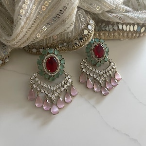May include: A pair of ornate earrings featuring a central oval red gemstone surrounded by small green stones. Below, a cascade of clear and pink teardrop-shaped gems hangs from a silver-toned setting. The earrings rest on a white surface with a decorative scarf.