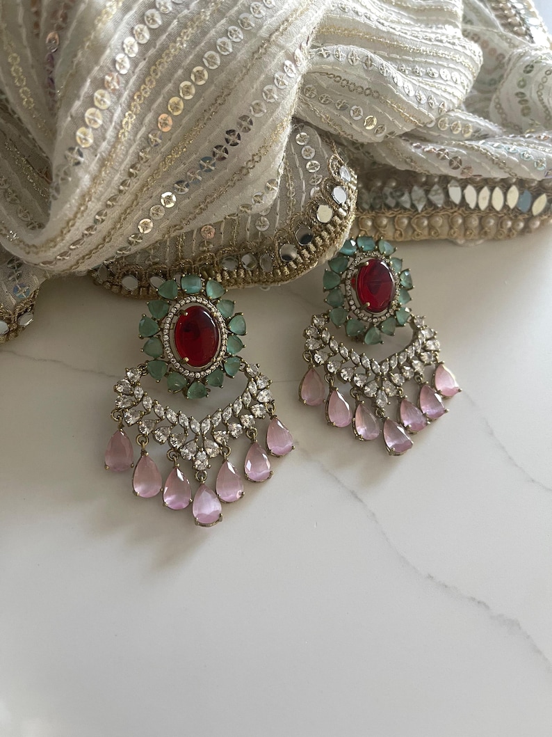 May include: A pair of ornate earrings featuring a central oval red gemstone surrounded by small green stones. Below, a cascade of clear and pink teardrop-shaped gems hangs from a silver-toned setting. The earrings rest on a white surface with a decorative scarf.