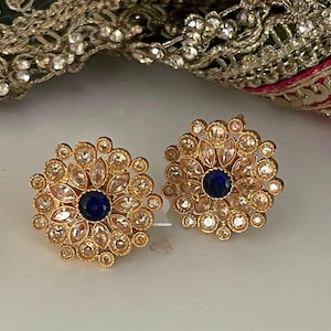 May include: A pair of gold-toned stud earrings. Each earring features a central dark blue stone surrounded by a ring of clear, oval-shaped stones. The earrings have a floral design and are set against a light background.