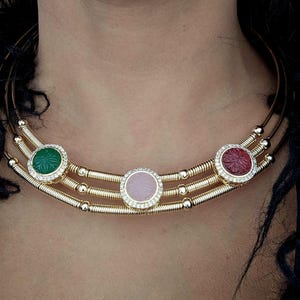 May include: A gold-toned necklace with three rows, featuring three circular gemstone pendants. The pendants are green, pink, and red, each surrounded by a ring of small, clear stones. The necklace is designed to sit around the neck.