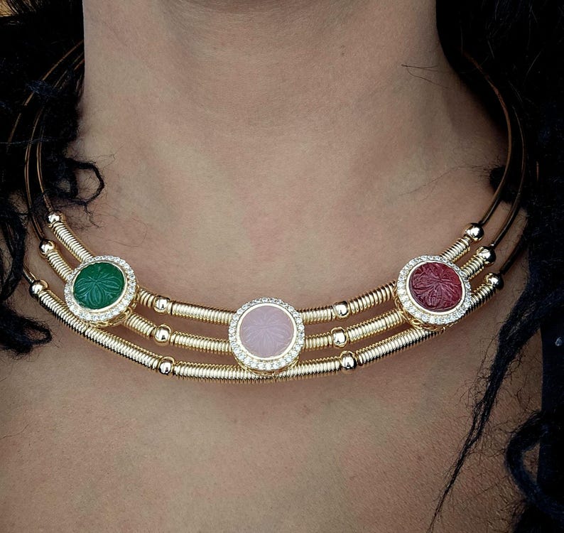 May include: A gold-toned necklace with three rows, featuring three circular gemstone pendants. The pendants are green, pink, and red, each surrounded by a ring of small, clear stones. The necklace is designed to sit around the neck.
