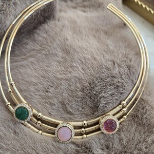 May include: A gold-toned collar necklace with three circular bands. The necklace features three gemstone pendants: green, pink, and purple, each surrounded by small clear stones. The necklace rests on a soft, gray surface.