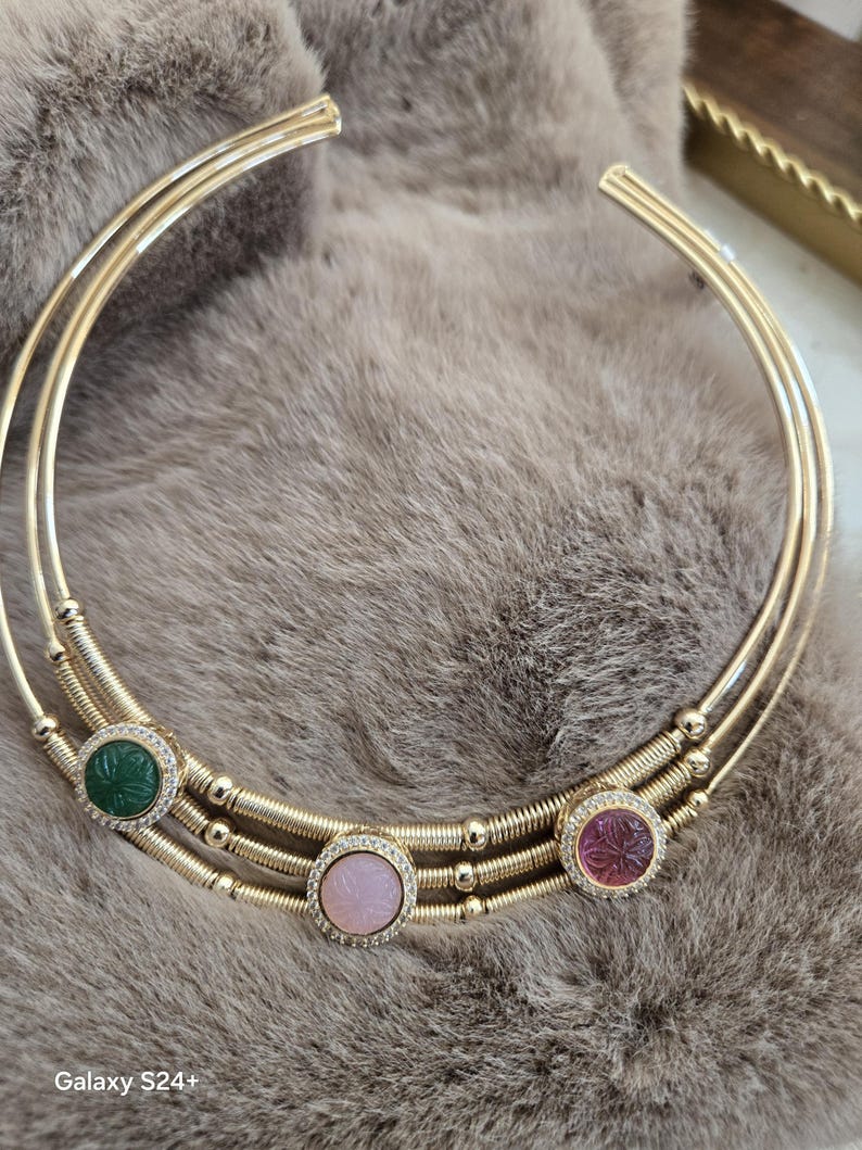May include: A gold-toned collar necklace with three circular bands. The necklace features three gemstone pendants: green, pink, and purple, each surrounded by small clear stones. The necklace rests on a soft, gray surface.