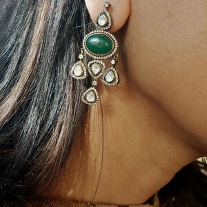 Puede incluir: Un par de pendientes colgantes con una piedra preciosa verde y brillantes transparentes. Los pendientes est&aacute;n hechos de metal dorado.