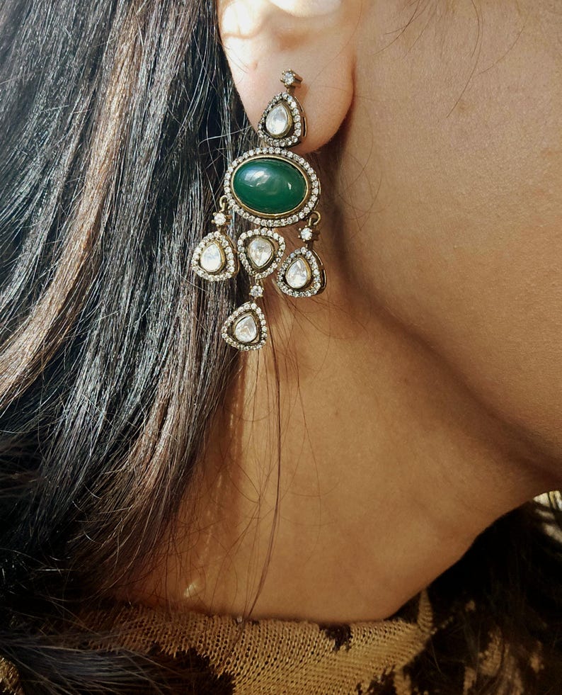 Puede incluir: Un par de pendientes colgantes con una piedra preciosa verde y brillantes transparentes. Los pendientes est&aacute;n hechos de metal dorado.