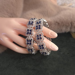 May include: Two silver bracelets with a floral design, each flower containing a dark blue gemstone. The bracelets are displayed on a mannequin hand against a dark green and tan background. The bracelets are a jewelry item.