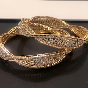 May include: Two gold-toned bangles featuring a twisted, openwork design. Each bangle is embellished with numerous small, clear, sparkling stones set along the intricate metal framework, creating a textured, reflective surface on the jewelry.