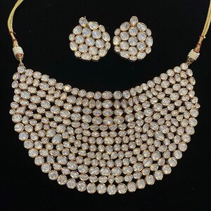 May include: A gold-coloured necklace and matching earrings set. The necklace has a bib design, densely covered with clear, oval-shaped stones. The earrings are round, also set with the same clear stones. The jewellery is displayed against a black background.