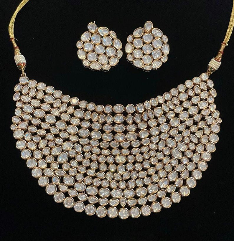 May include: A gold-coloured necklace and matching earrings set. The necklace has a bib design, densely covered with clear, oval-shaped stones. The earrings are round, also set with the same clear stones. The jewellery is displayed against a black background.