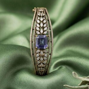 May include: An ornate gold-toned bracelet with a large, square, blue-purple gemstone at its center. The bracelet is encrusted with small, clear gemstones and features a leaf-like design. The background is a draped, green fabric.