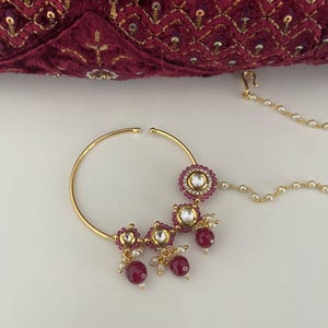 May include: A gold-colored nose ring with three decorative clusters of red gemstones and white pearls. The nose ring has a large circular hoop and a delicate chain with small white pearls. The background features a burgundy velvet fabric.