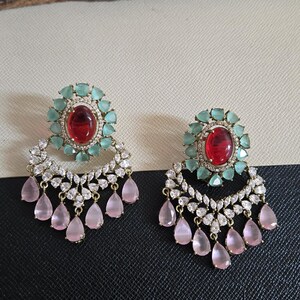 May include: A pair of ornate earrings featuring a central red gemstone surrounded by light green stones. Below, a cascade of clear and pink teardrop-shaped gems hangs from a gold-colored setting. The earrings are displayed on a black and white surface.