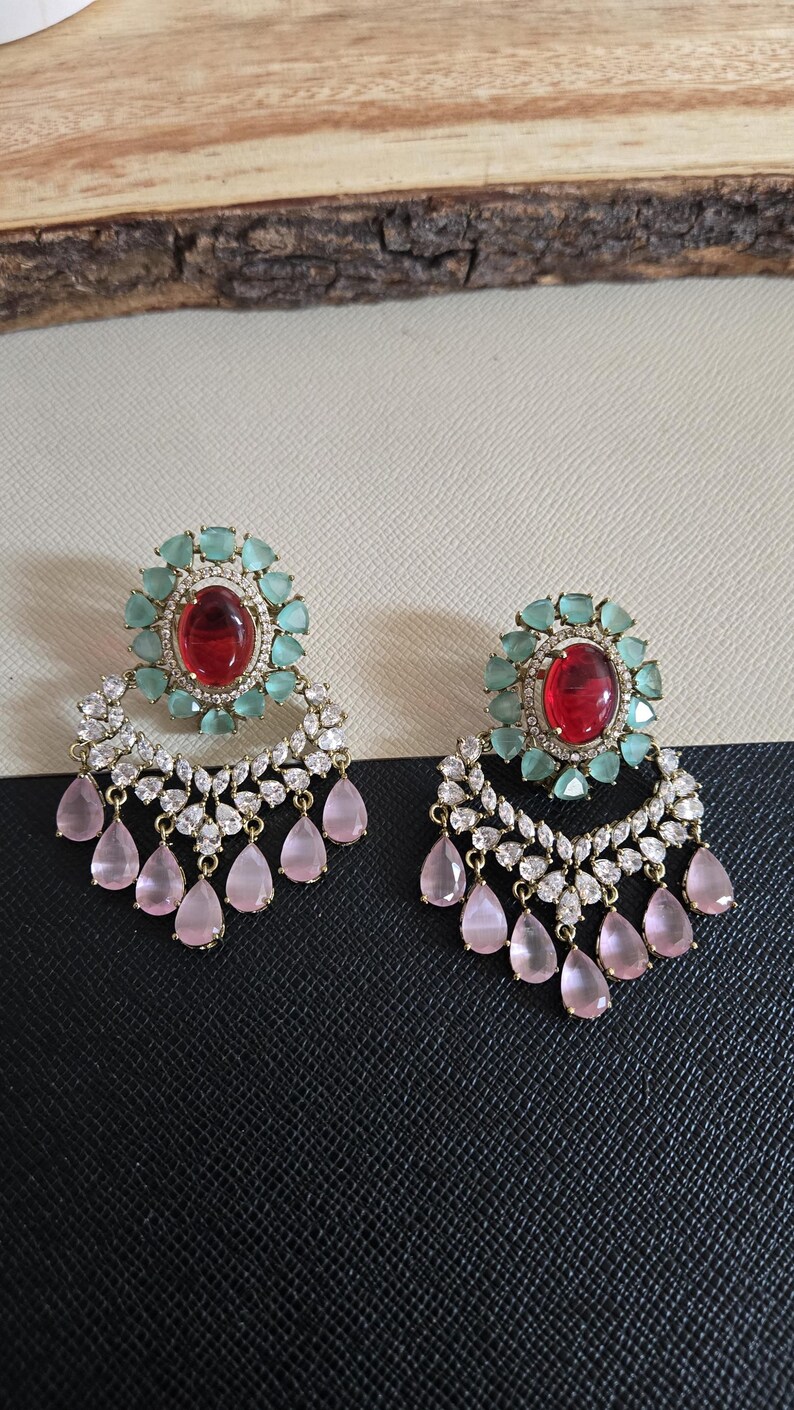 May include: A pair of ornate earrings featuring a central red gemstone surrounded by light green stones. Below, a cascade of clear and pink teardrop-shaped gems hangs from a gold-colored setting. The earrings are displayed on a black and white surface.