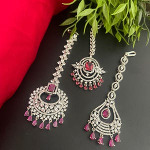 May include: Three-piece jewelry set featuring silver-toned metal and sparkling clear and ruby-colored stones. The set includes a headpiece and two earrings, all with intricate designs and dangling ruby-colored accents. The jewelry is displayed on a black surface.