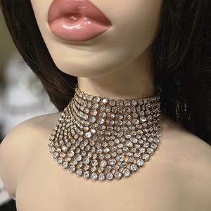 May include: A gold choker necklace set with numerous clear, round gemstones. The necklace is displayed on a mannequin, highlighting its design and sparkle. The gemstones are closely set, creating a dense, glittering effect.