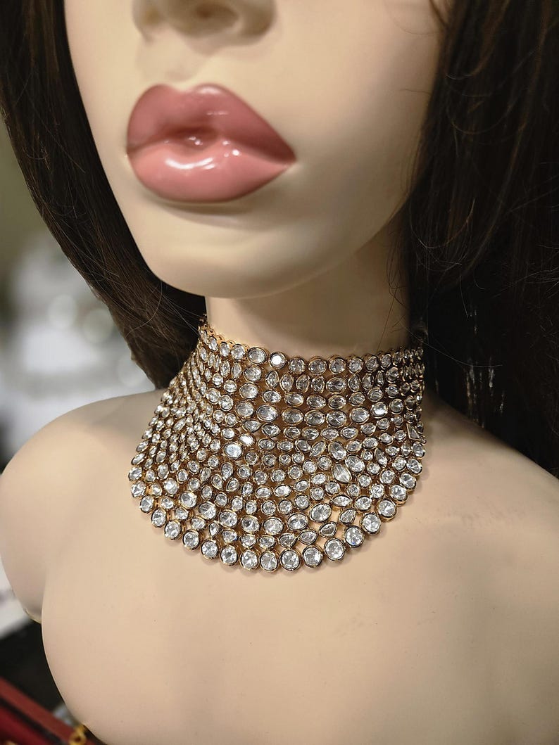 May include: A gold choker necklace set with numerous clear, round gemstones. The necklace is displayed on a mannequin, highlighting its design and sparkle. The gemstones are closely set, creating a dense, glittering effect.