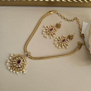May include: A gold-coloured necklace and earring set. The necklace has a pendant with a central red gemstone surrounded by small white pearls and clear crystals. The matching earrings have a similar design. The jewellery is displayed on a light-coloured surface.