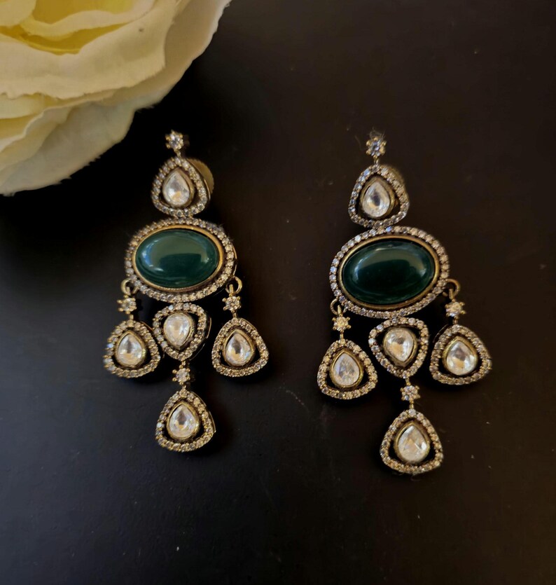 Puede incluir: Un par de pendientes colgantes con una piedra verde y brillantes transparentes. Los pendientes son de color dorado y tienen forma de l&aacute;grima.