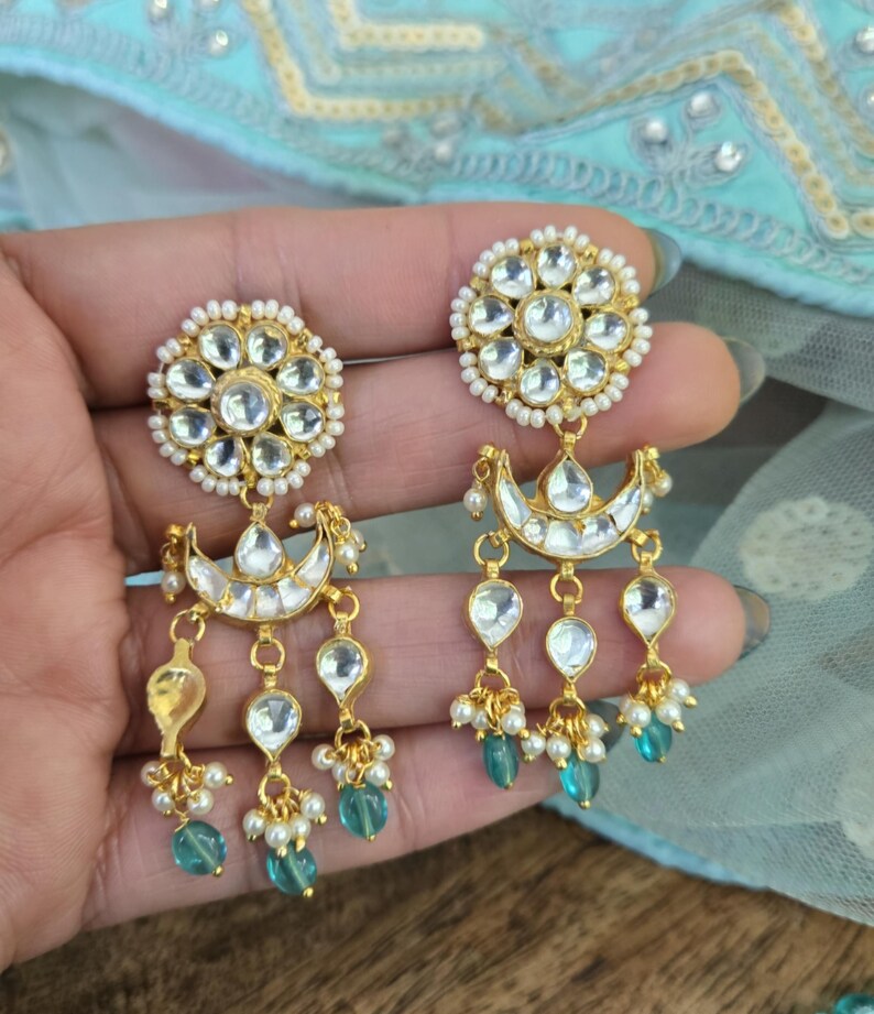 Op de afbeelding: Gouden oorbellen met een bloemmotief en traanvormige accenten. De oorbellen hebben een halve maanvorm en zijn versierd met witte parels en turquoise blauwe stenen.