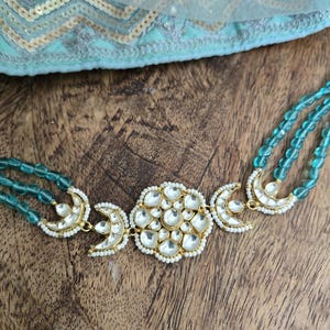 Op de afbeelding: Een gouden ketting met een grote, sierlijke hanger met een bloemmotief met witte stenen. De ketting is gemaakt van drie strengen turquoise kralen.