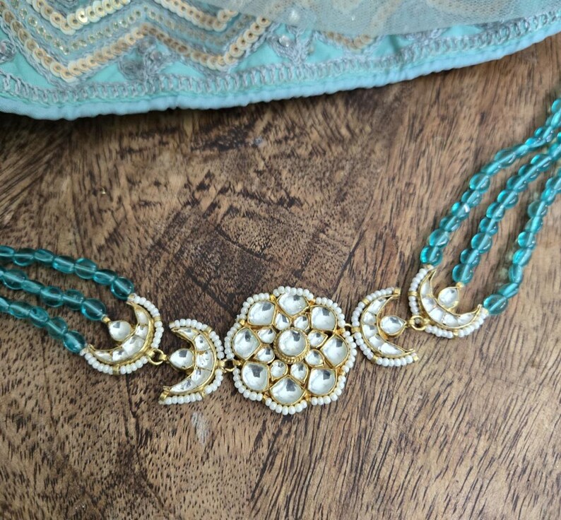 Op de afbeelding: Een gouden ketting met een grote, sierlijke hanger met een bloemmotief met witte stenen. De ketting is gemaakt van drie strengen turquoise kralen.