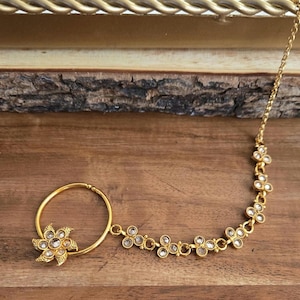 May include: A gold-colored nose ring with a floral design and a chain of small flower-shaped accents. The nose ring features a large, flower-shaped embellishment with clear, sparkling stones. The chain extends to a series of delicate flower-shaped links.